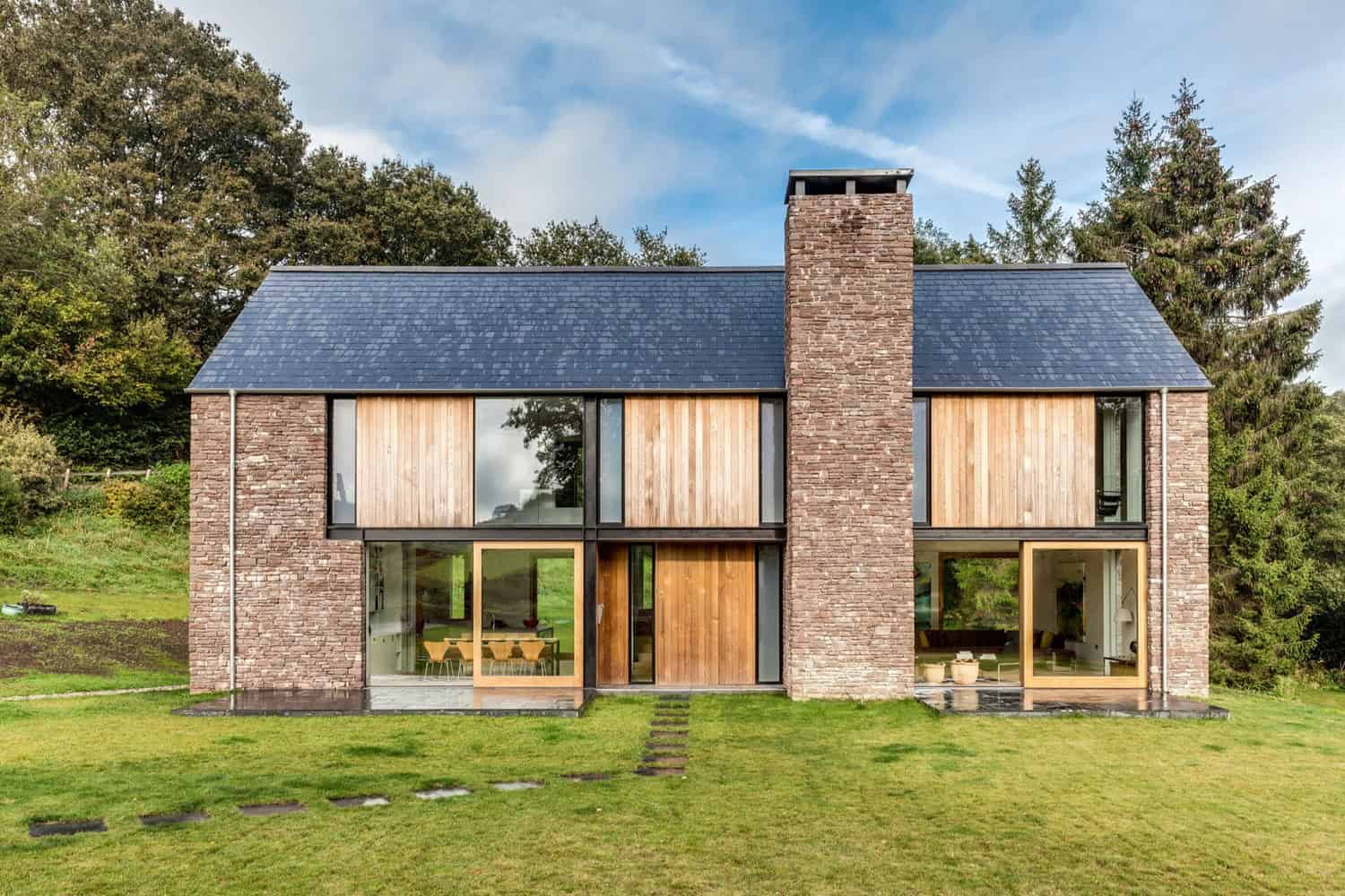 Modern brick and wood house with large glass windows and a sloped roof, surrounded by lush greenery and trees, showcasing contemporary architectural design and innovative use of natural materials.
