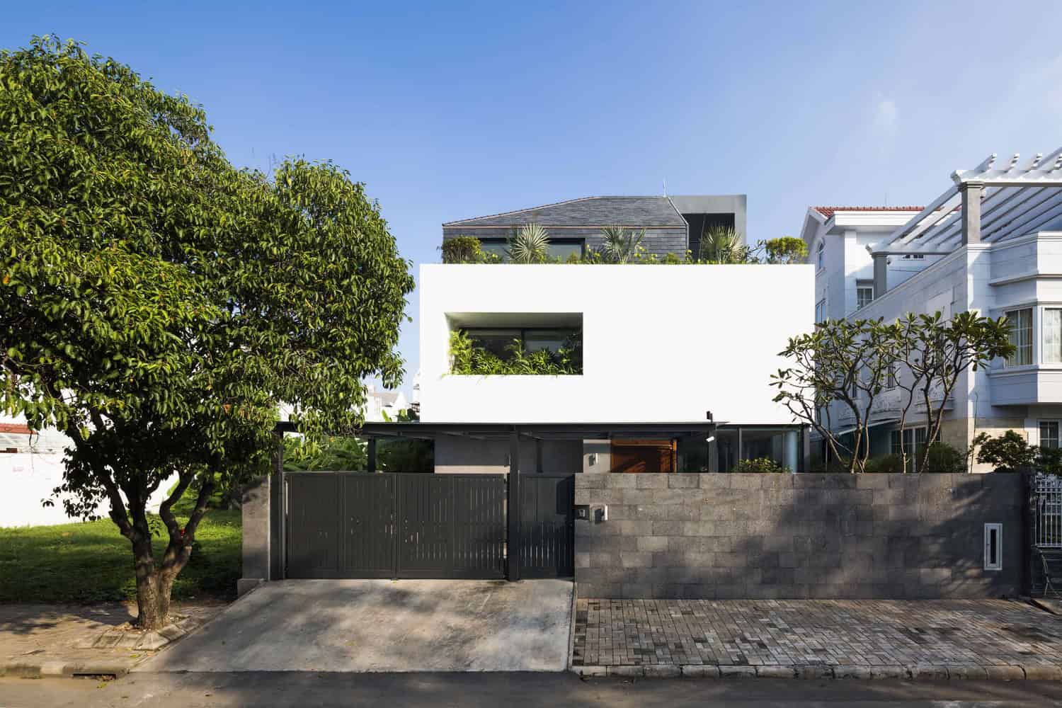 Contemporary minimalist modern white concrete house with clean lines, large window, lush greenery, and a sleek black gate in an urban residential neighborhood.