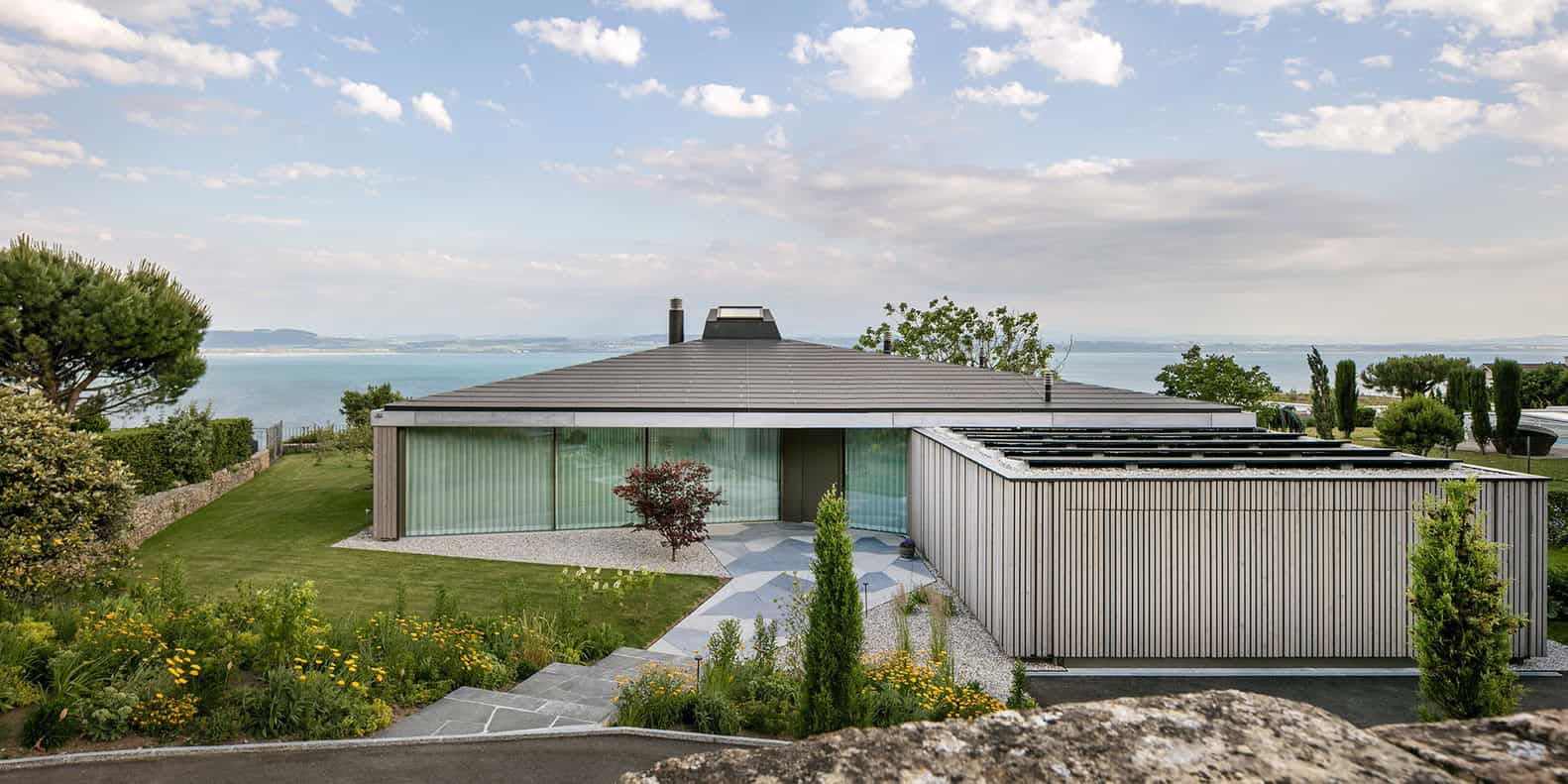 Contemporary modern house with minimalist design, large glass windows, and natural wood siding overlooking a scenic water view, surrounded by lush landscaping and vibrant garden.