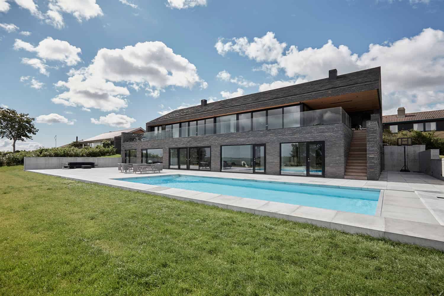 Contemporary modern house with black brick exterior, large glass windows, outdoor pool, and spacious backyard under a bright blue sky.