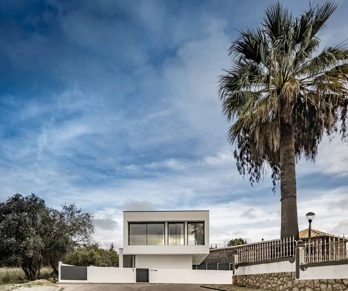 Modern minimalist house with white facade and large glass windows in a scenic landscape with a tall palm tree and clear blue sky.