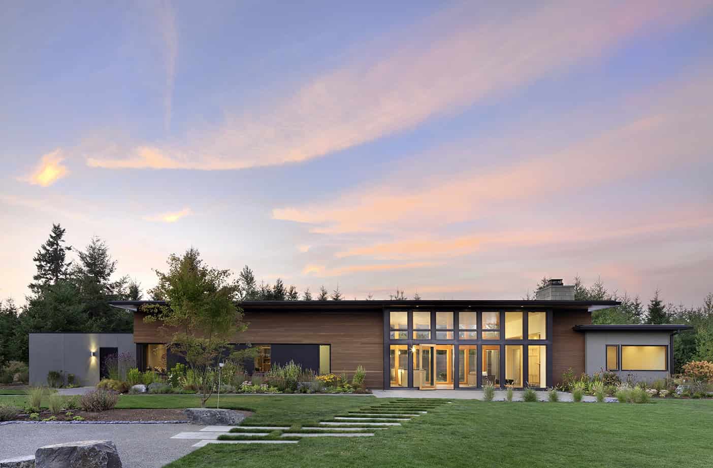 Contemporary modern house with large glass windows, wooden siding, and landscaped yard during sunset, showcasing minimalist architectural design and eco-friendly outdoor landscaping.