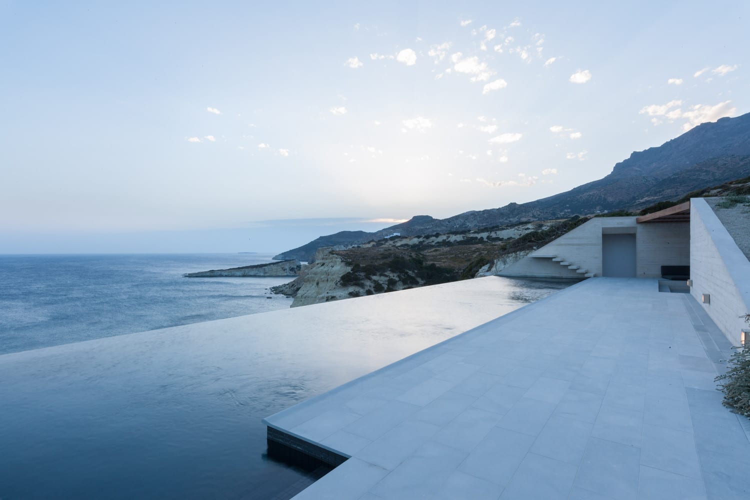 Seaside modern minimalist home with infinity pool overlooking ocean cliffs and mountain landscape. Contemporary architecture blending natural views and sleek design elements.