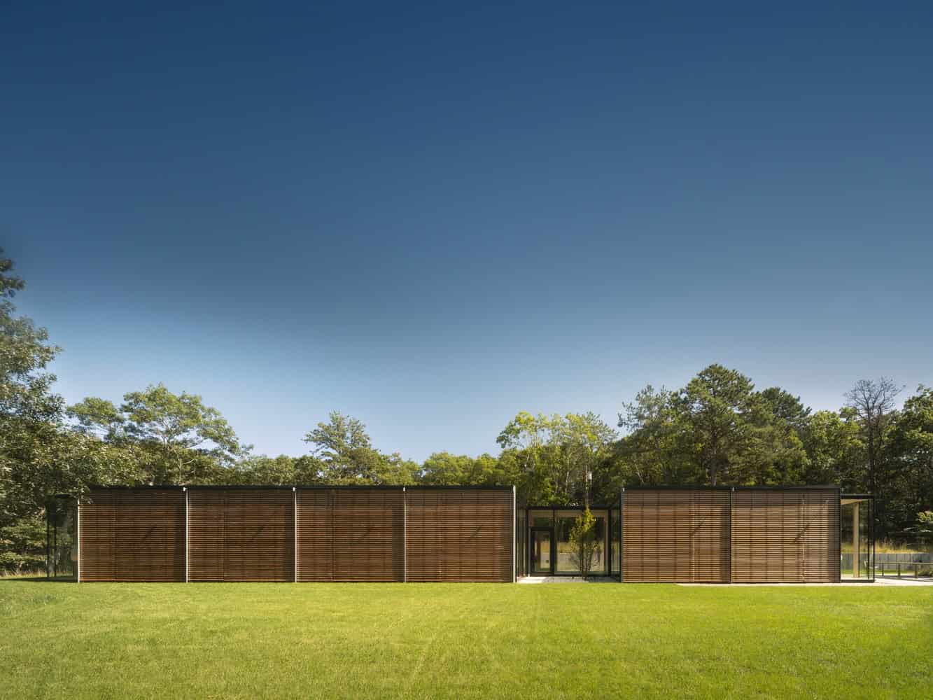 Modern minimalist wooden house with sleek lines and large glass windows, set amidst lush greenery and open lawn, exemplifying contemporary architecture and innovative design.