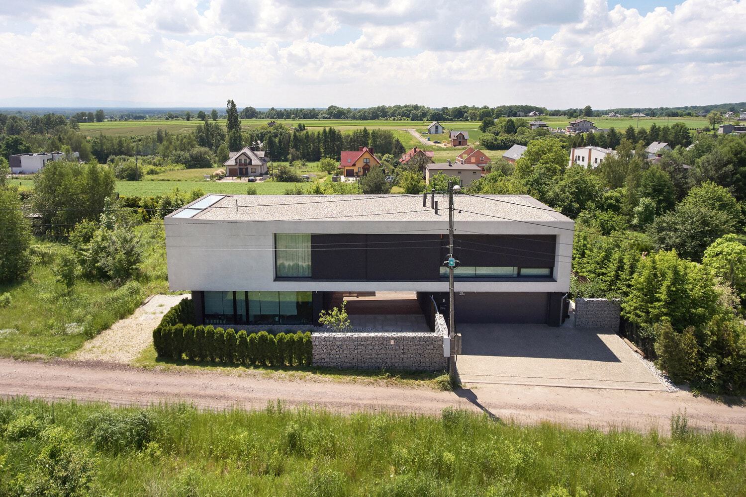 Modern minimalist house with sleek black and white exterior featuring large glass windows and a spacious driveway, surrounded by lush greenery and scenic rural landscape.
