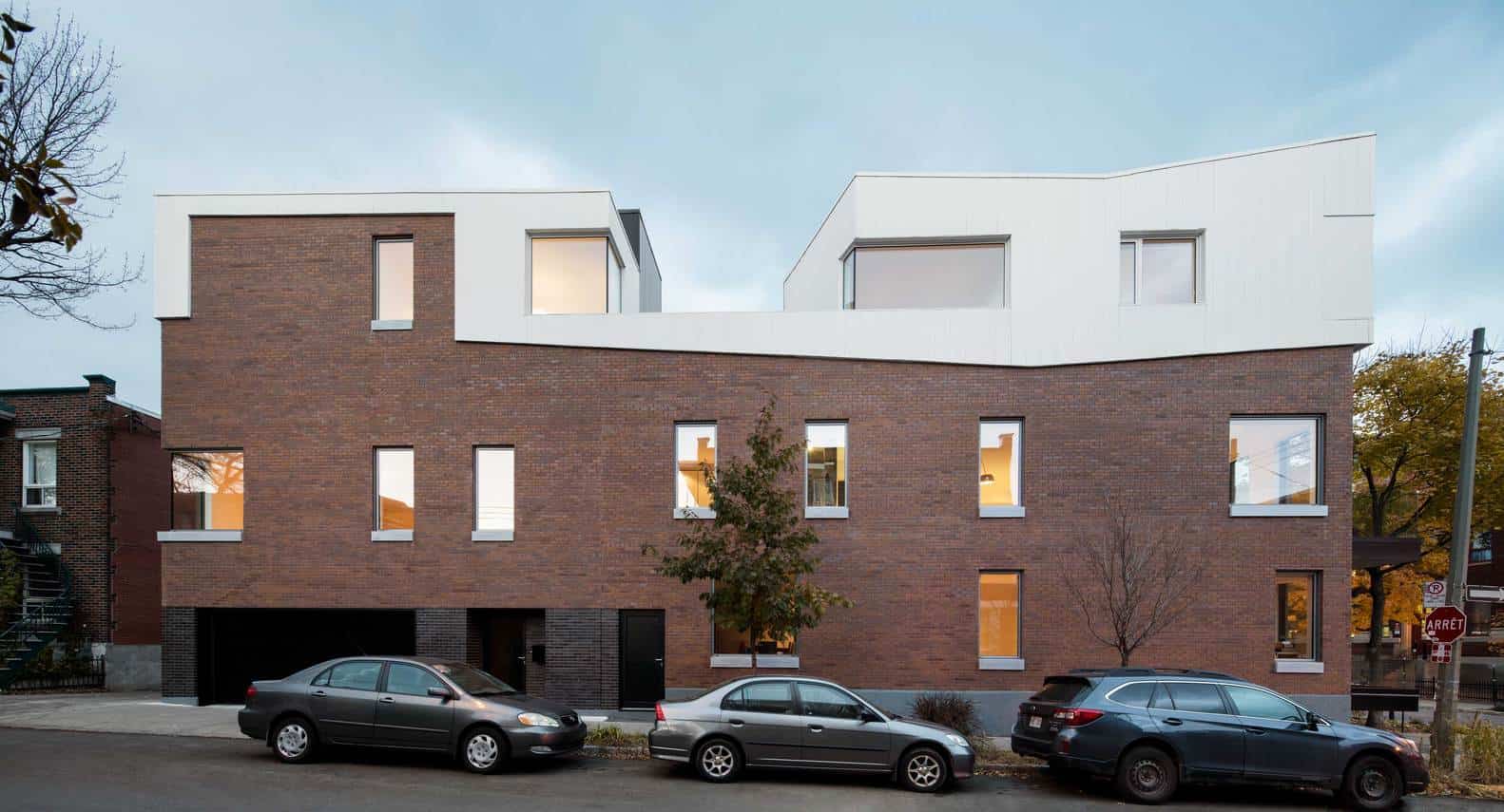 Contemporary modern residential building with brick facade and white geometric upper structures, surrounded by parked cars in an urban setting.