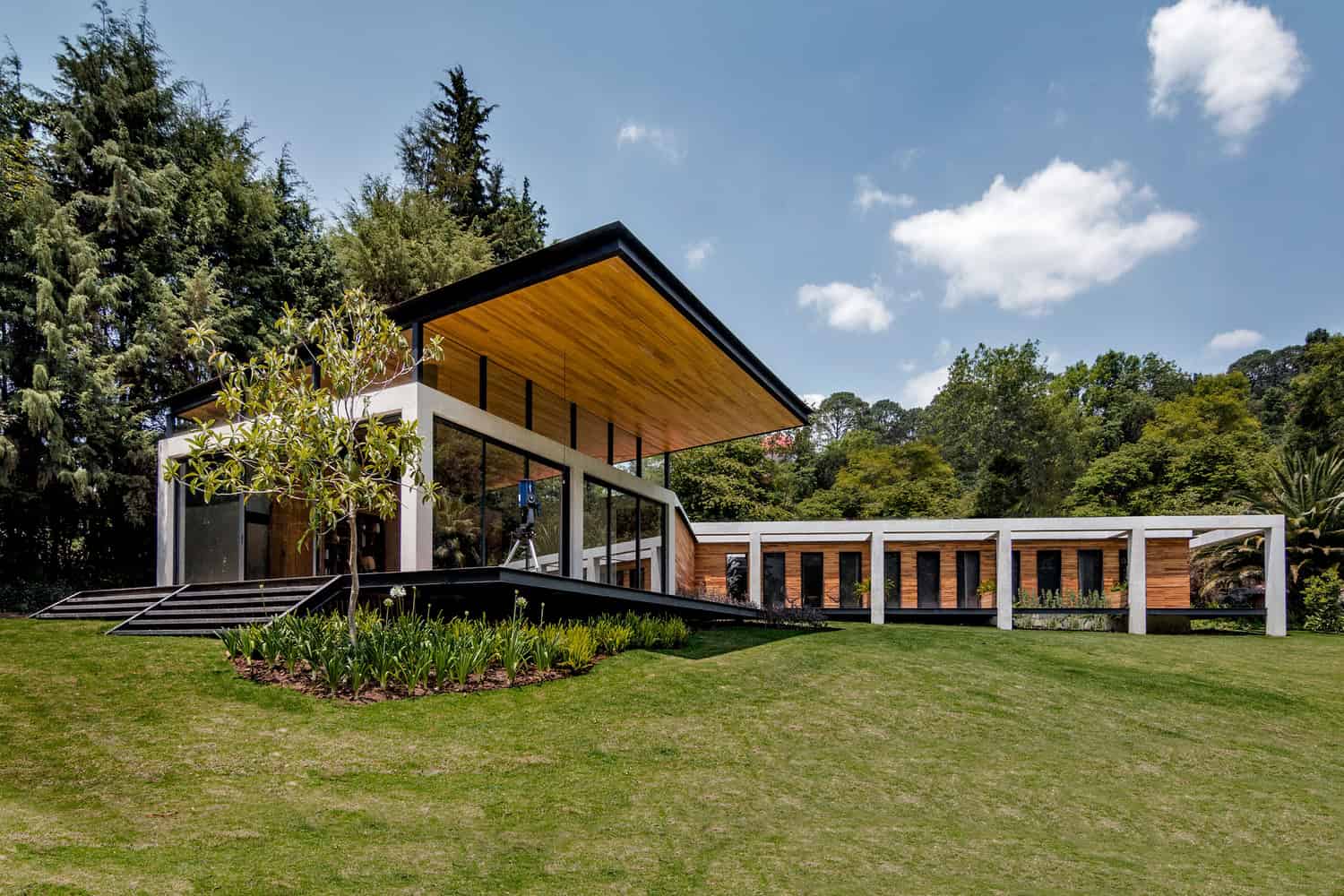 Contemporary modern house with large glass windows, wooden accents, and an overhanging roof, nestled in lush greenery on a sloped landscape, showcasing innovative architecture and outdoor design.