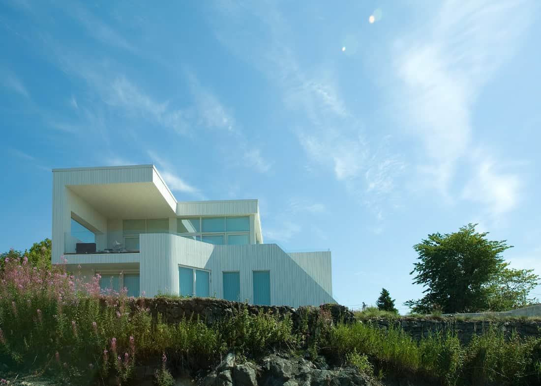 Modern minimalist white house with large glass windows on a hilltop under a bright blue sky.