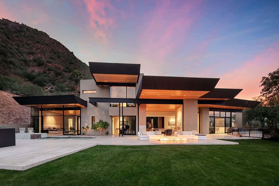 Contemporary luxury modern house with sleek design, large glass windows, and outdoor fire pit, set against a scenic mountain backdrop at sunset, perfect for upscale residential architecture.