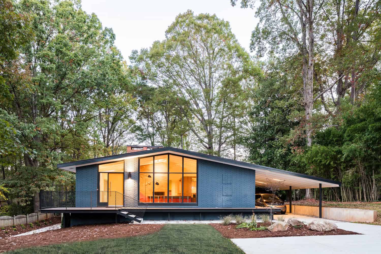 Modern mid-century style house with large glass windows and a sloped roof, surrounded by lush trees and landscaped yard, exemplifying innovative architectural design and home exterior elegance.