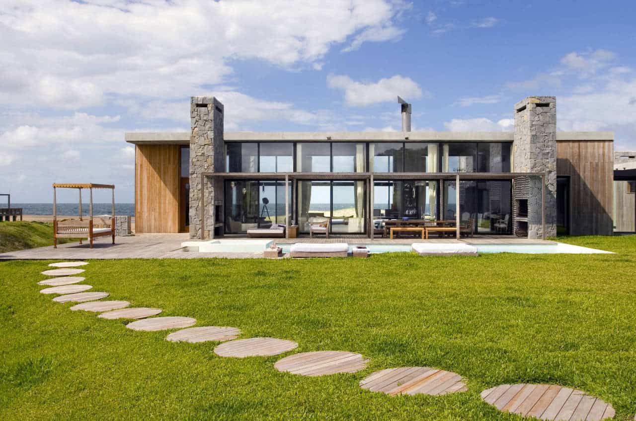 Modern beachfront house with large glass windows, stone accents, wooden features, outdoor seating, and lush green lawn. A stunning example of contemporary coastal architecture and design.