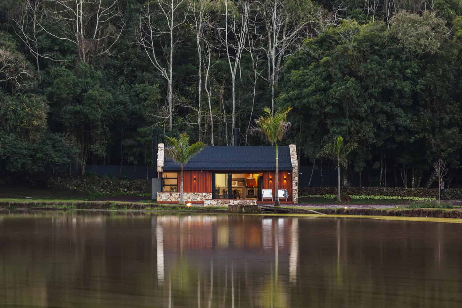 Modern lakeside tiny house with wood and stone exterior, surrounded by lush forest and palm trees, reflecting on tranquil water, ideal for eco-friendly living and outdoor relaxation.