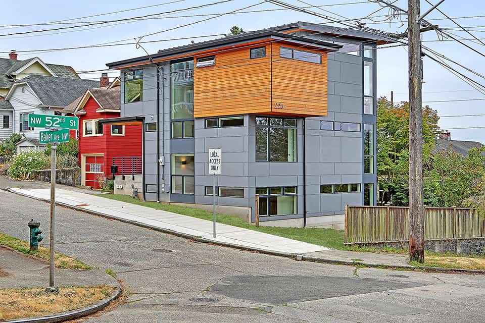 Modern urban residential building with sleek design, gray and wood exterior, large windows, located on a corner lot in a neighborhood.