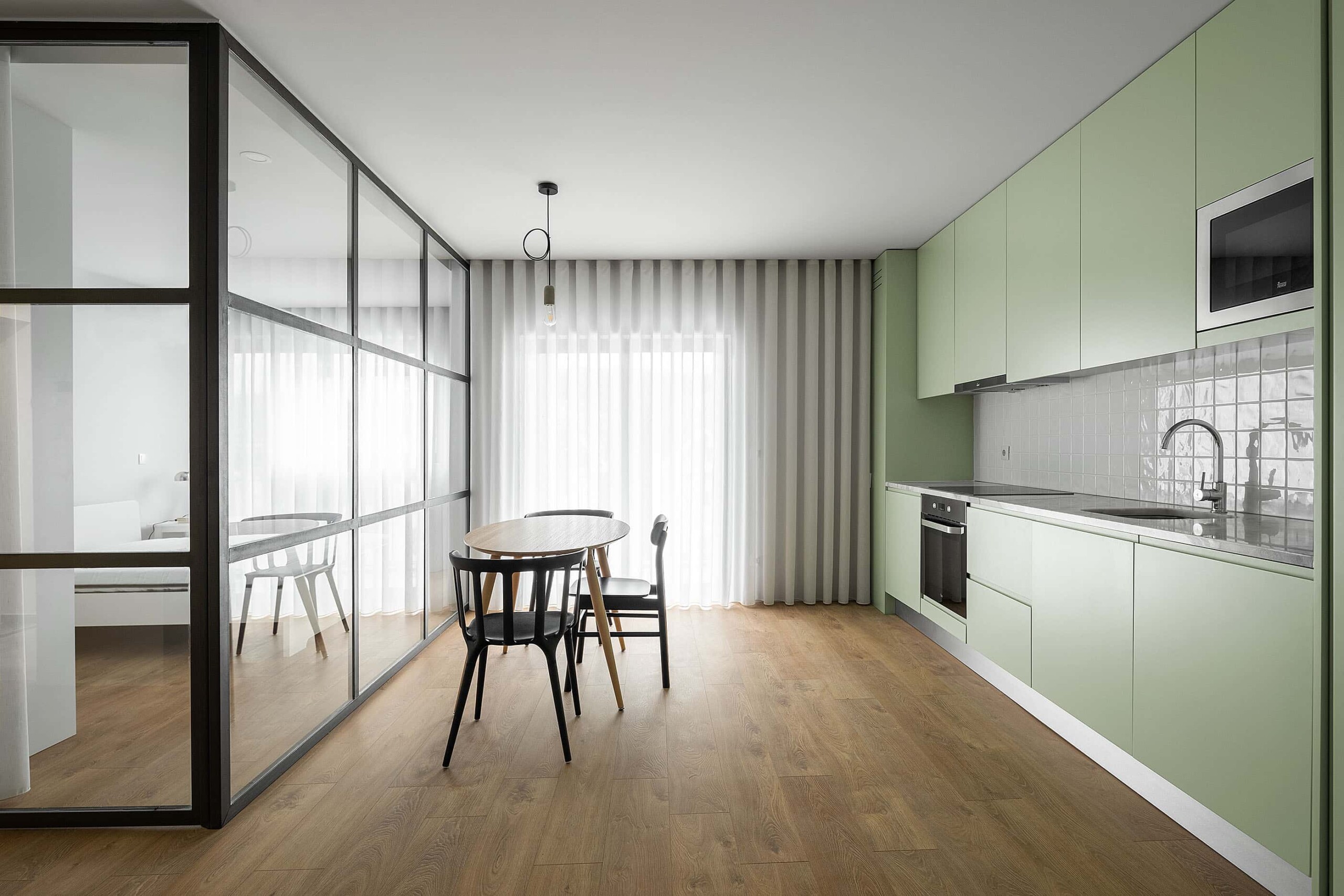 Modern minimalist kitchen with light green cabinetry and open-concept design, featuring wood flooring, sleek appliances, and a glass partition separating dining and living areas.
