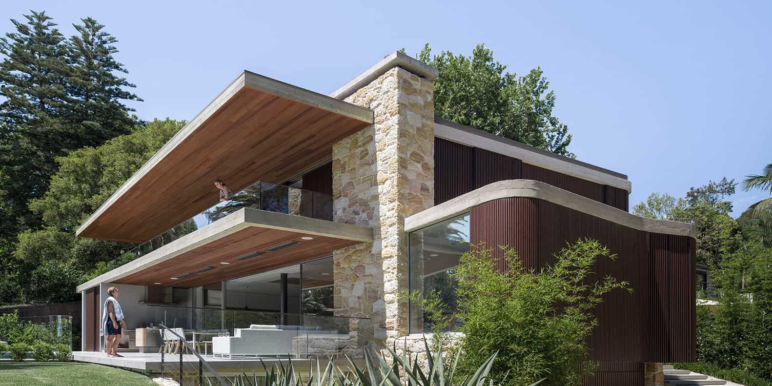 Contemporary modern house with natural stone and wood accents, large glass windows, and sleek architectural design surrounded by lush greenery.