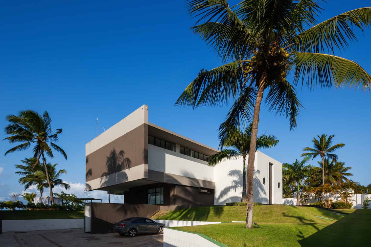 Luxurious modern hillside house with minimalist design, geometric architecture, and tropical palm trees under a clear blue sky.