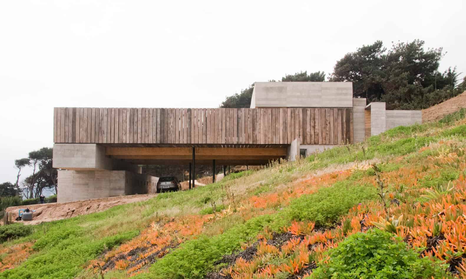 Sleek modern house with minimalist architecture, featuring concrete and wood elements on a hillside with lush greenery and colorful ground cover.