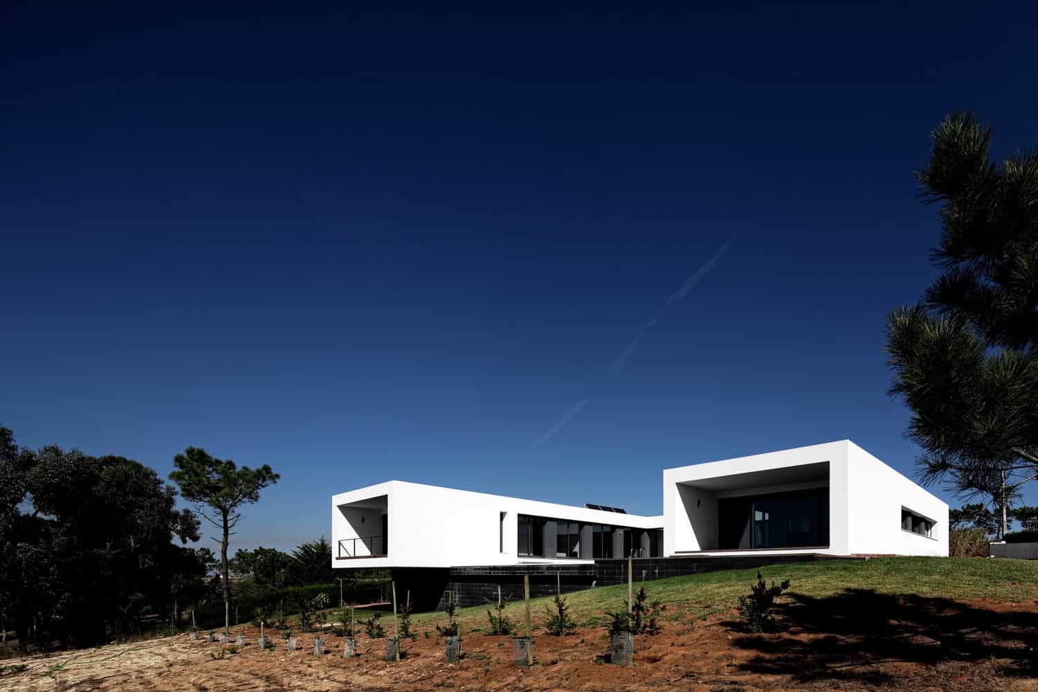 Modern minimalist white house with geometric design and large windows on grassy landscape under a clear blue sky.