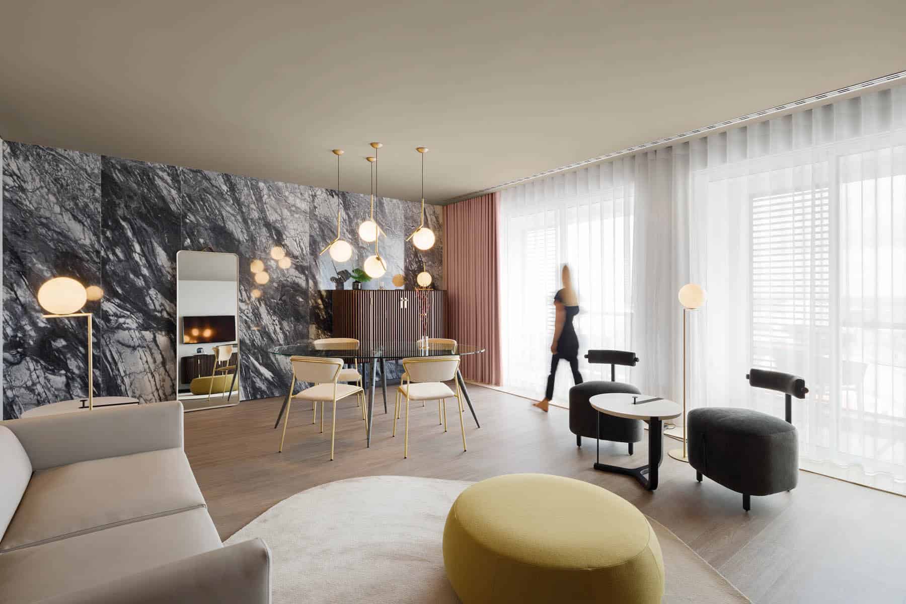 Modern living room interior with stylish furniture, marble accent wall, and large windows with sheer curtains.