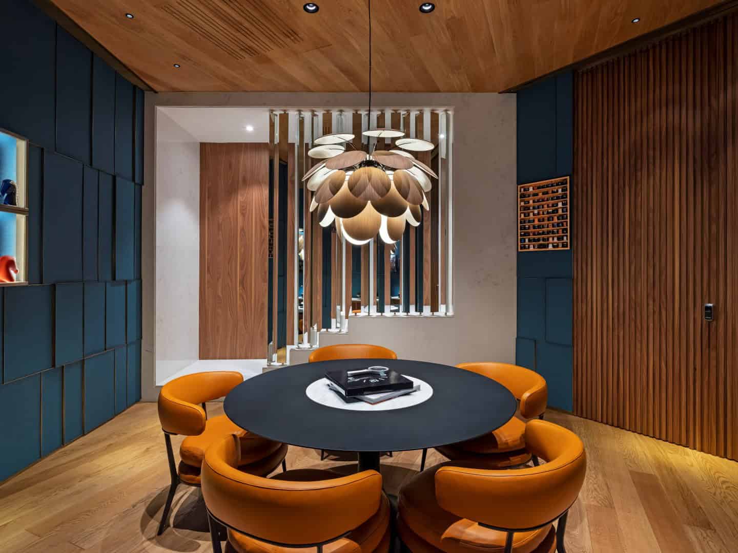 Contemporary dining room with custom wooden wall panels, stylish orange chairs, modern pendant lighting, and a round black table, showcasing modern interior design and architectural elegance.