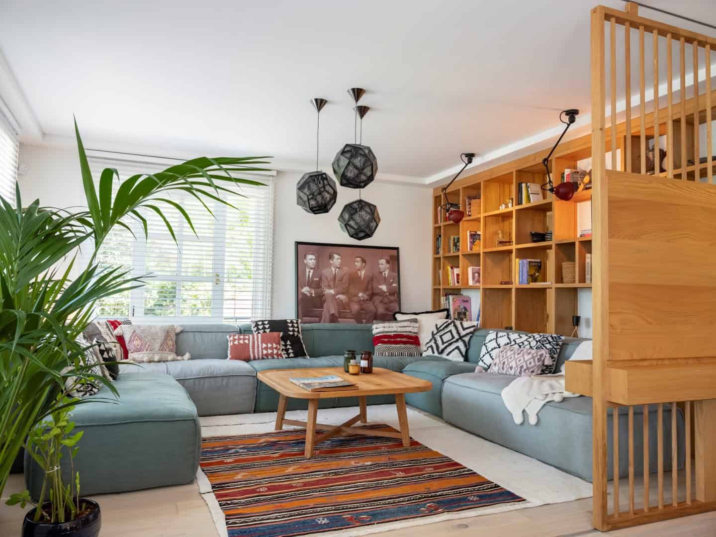 Bright contemporary living room with large windows, modern sectional sofa, colorful patterned cushions, wooden coffee table, and a spacious wooden bookshelf with wall-mounted lamps, showcasing stylish interior design.