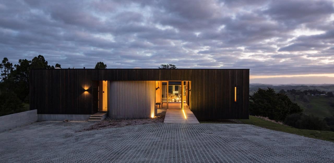 Modern minimalist black house with warm interior lighting, situated on a hillside at sunset, showcasing contemporary architecture and scenic landscape views.