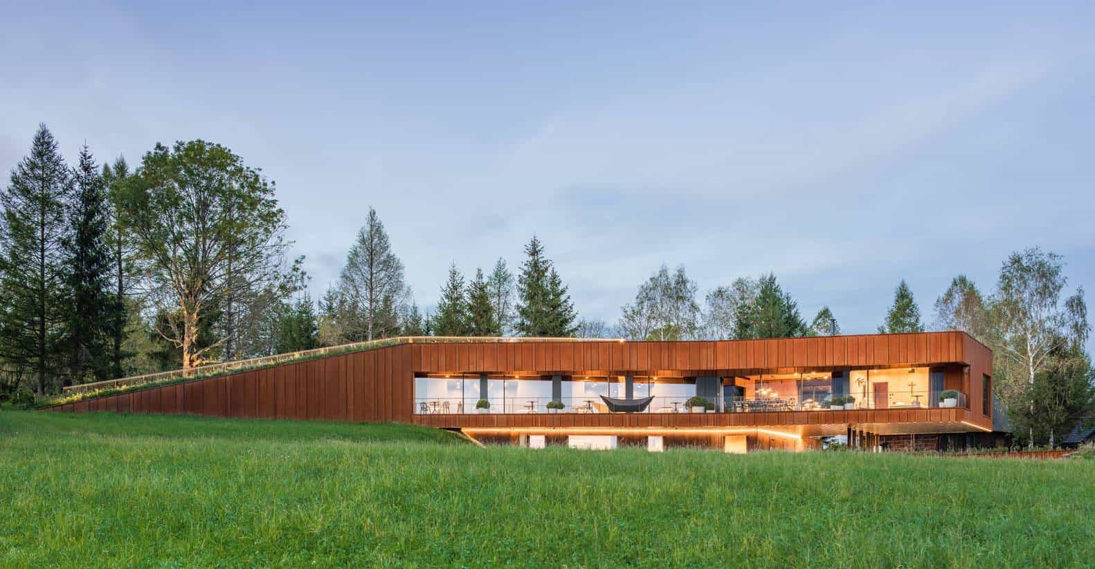 Modern eco-friendly house with curved wooden facade and large glass windows surrounded by greenery and trees.