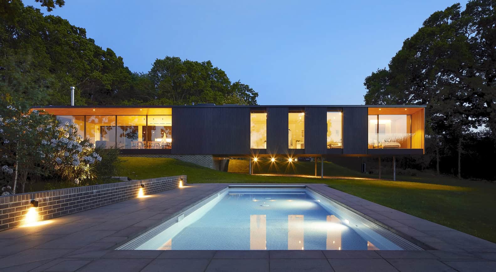 Contemporary modern house with sleek black exterior and large windows, illuminated at dusk, featuring a swimming pool and landscaped yard, showcasing innovative architectural design and stylish outdoor living.