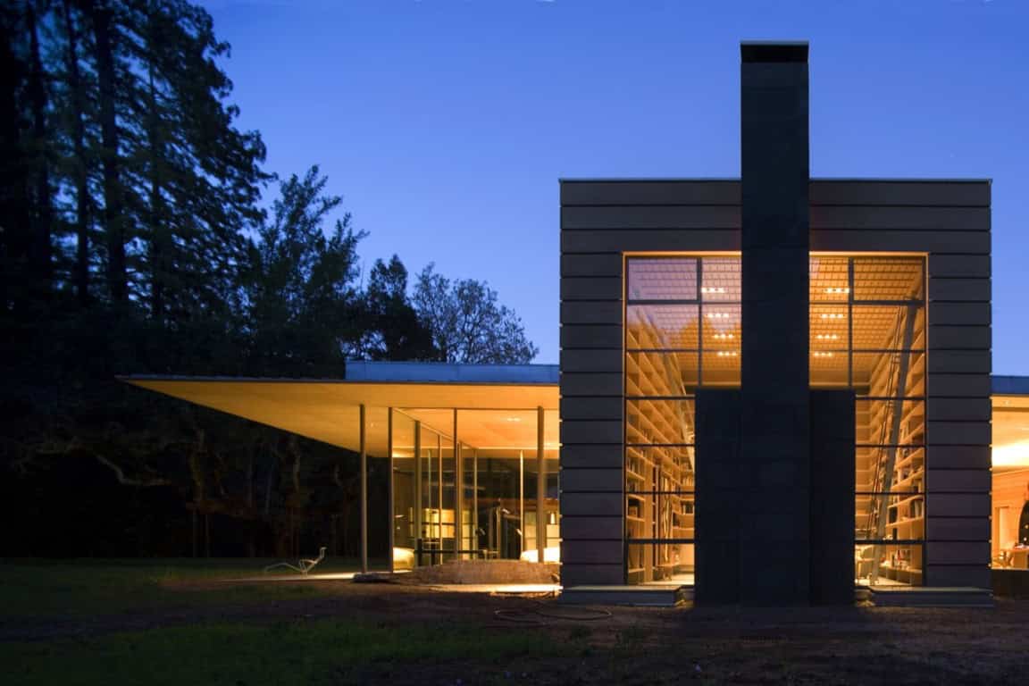 Futuristic modern house with large glass windows and sleek architectural design illuminated at dusk in a wooded environment.
