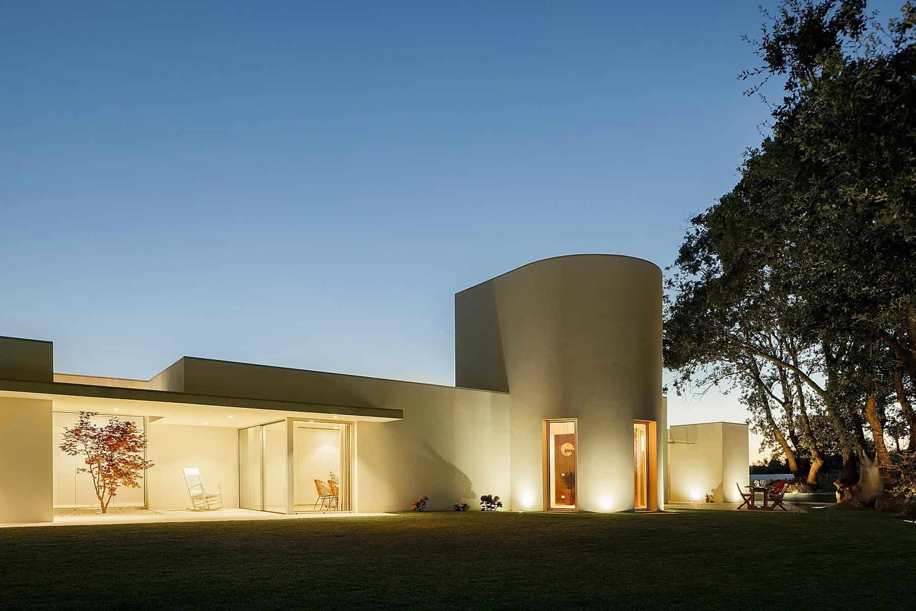 Modern minimalist house with curved and angular architectural design, illuminated outdoor lighting, and surrounding greenery in a tranquil evening setting.
