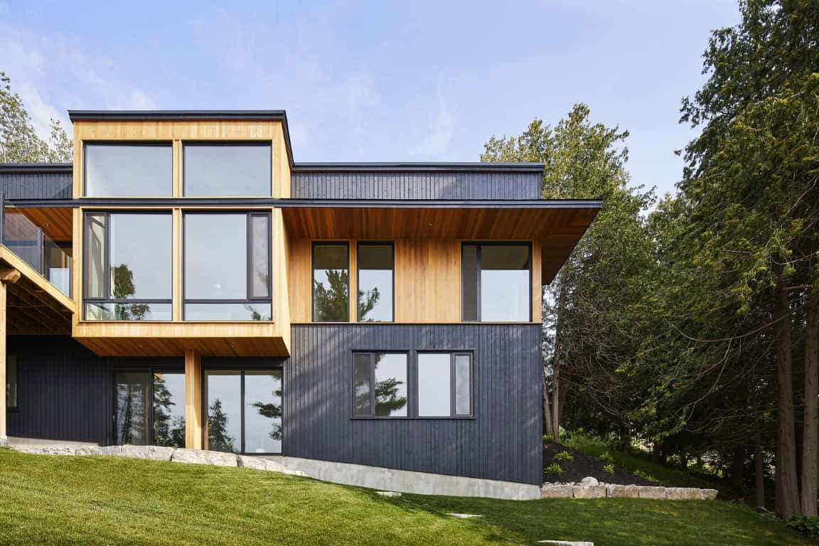 Contemporary modern house with black and wooden exterior design, large floor-to-ceiling windows, situated on a grassy sloped landscape surrounded by trees, showcasing innovative architecture.