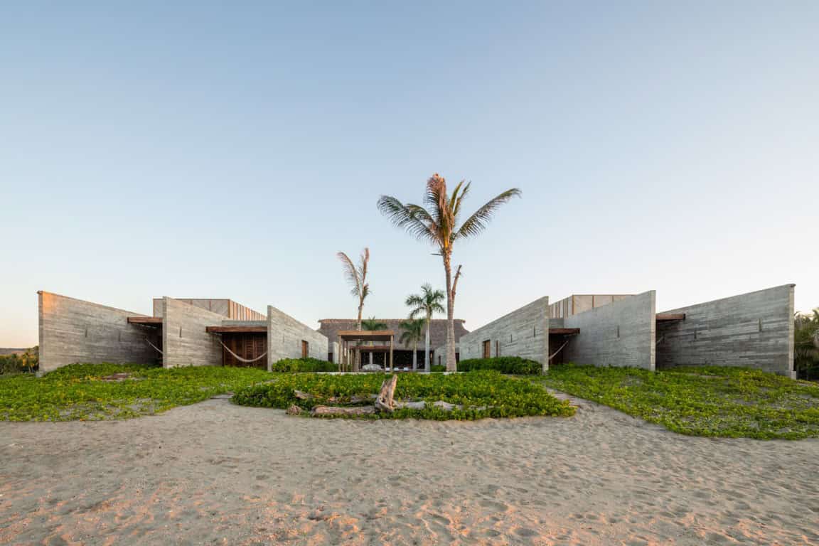 Modern beach house with contemporary architecture, concrete walls, tropical landscaping, and palm trees on sandy shoreline.