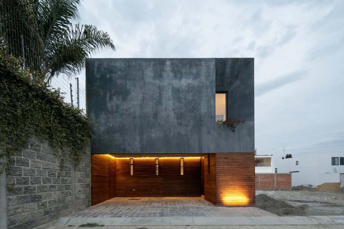 Contemporary minimalist house with a modern concrete facade and warm illuminated wooden garage entrance, set against a cloudy sky, showcasing sleek architectural design and innovative urban housing.