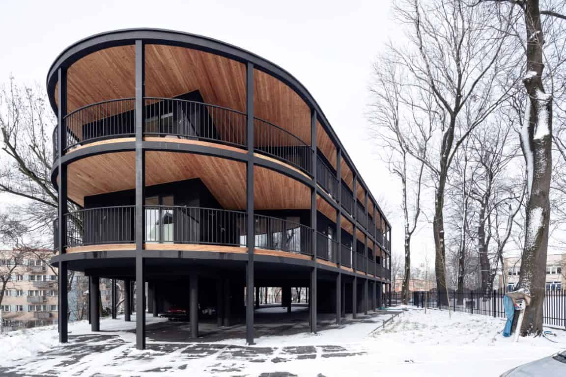 Modern multi-story residential building with curved wooden balconies and black metal framing, located in a snowy urban landscape during winter.