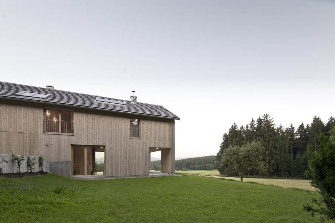 Modern wooden house with minimalist architecture in a scenic countryside setting, featuring large windows and a lush green lawn, emphasizing eco-friendly design and natural materials.