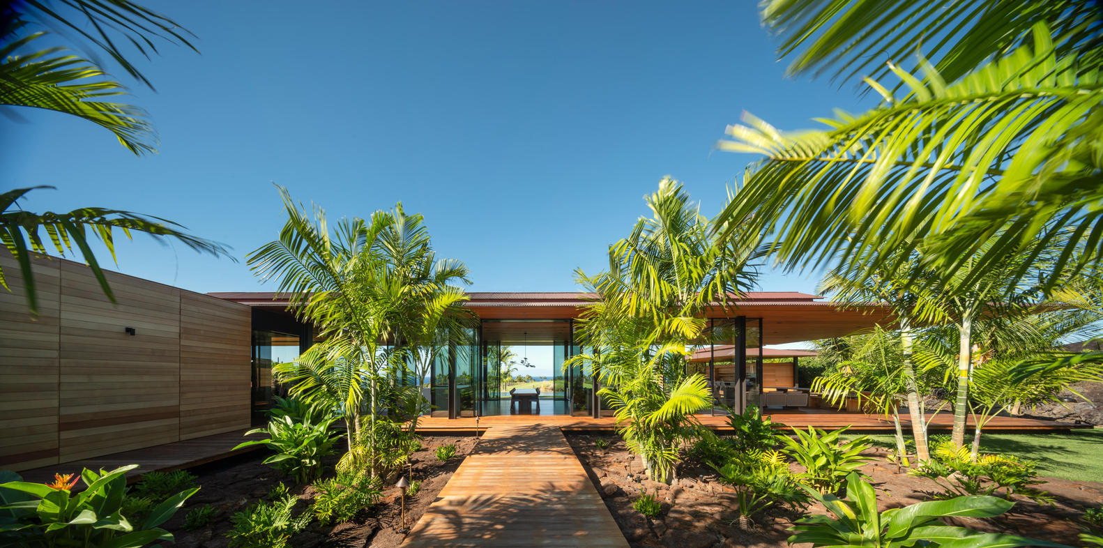Modern tropical house with outdoor wooden deck, lush green palm trees, and glass walls, surrounded by vibrant garden, under clear blue sky.