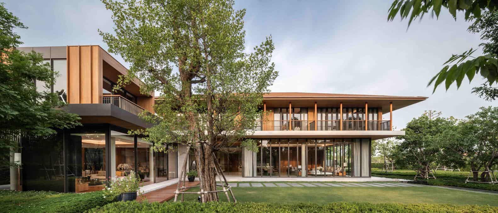 Modern two-story house with large glass windows and wooden accents surrounded by greenery and landscaped garden.