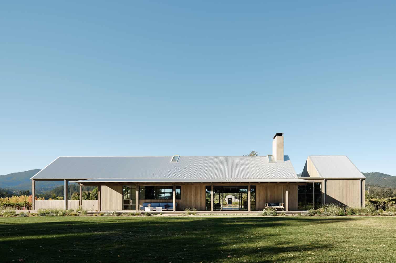 Modern rural house with wooden exterior and large glass windows under a clear blue sky.