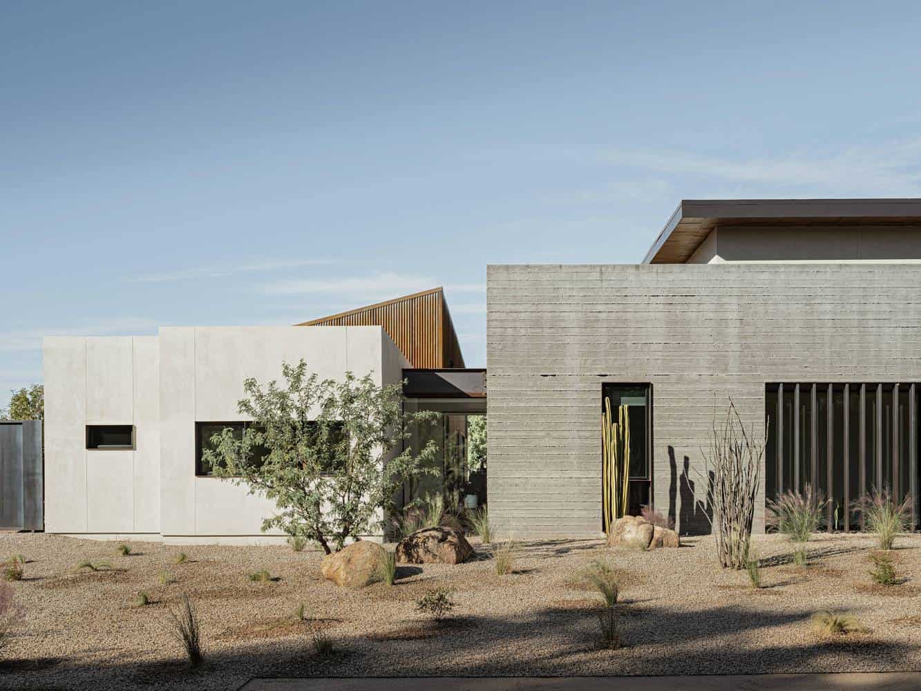 Contemporary desert-style house with minimalist architecture, featuring concrete and wood exterior design, large windows, and desert landscaping with rocks and drought-tolerant plants.