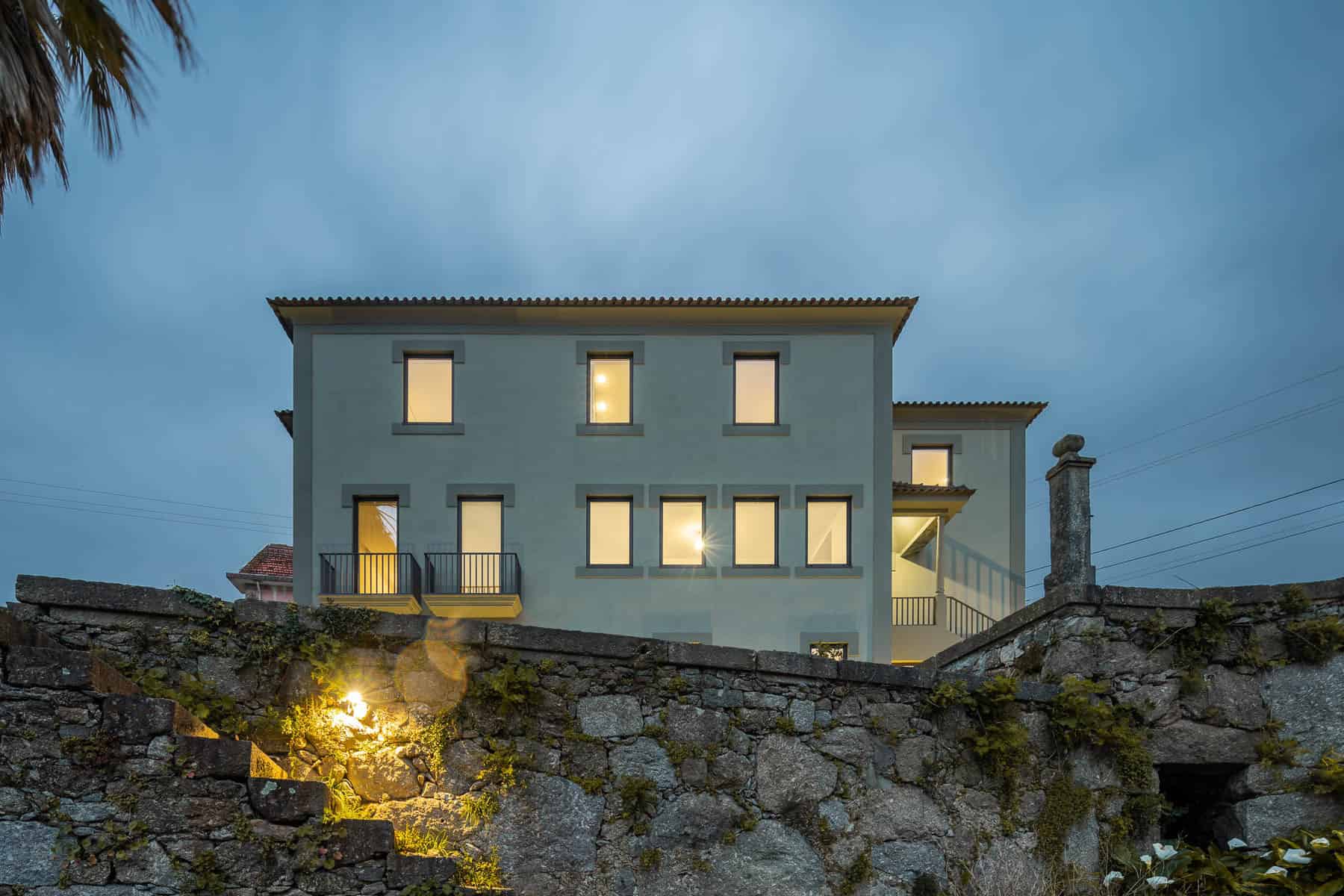 Contemporary modern house with large illuminated windows and balcony, situated above an old stone wall, showcasing stylish architecture with a peaceful evening sky background.