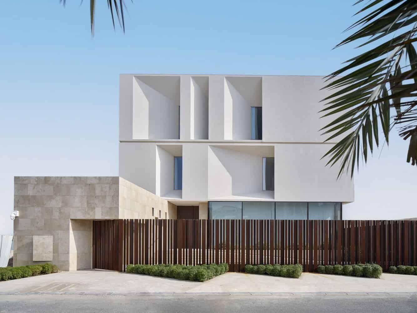 Modern minimalist residential building with geometric white facade and innovative window design, surrounded by landscaped greenery and wooden fencing.