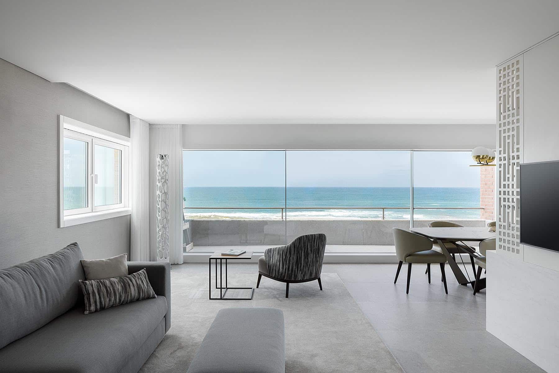 Modern ocean-view apartment living room with large glass balcony overlooking the beach, minimalist furniture, and neutral color palette, showcasing contemporary architecture and interior design.