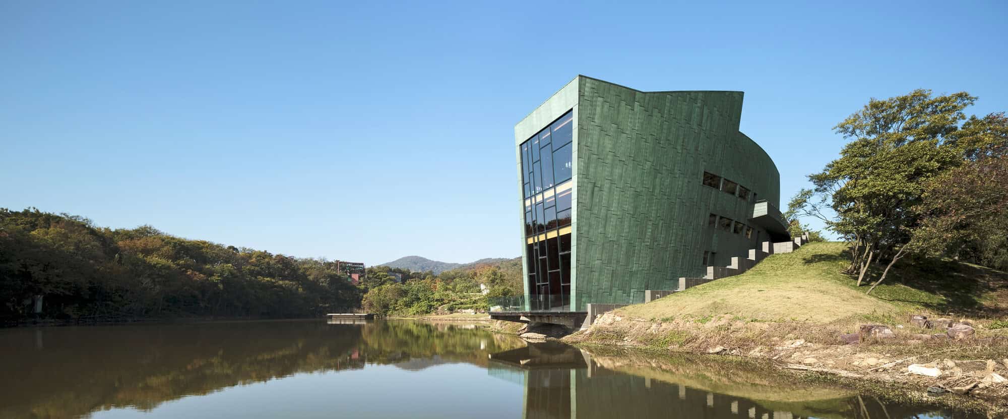 Modern green architectural building with reflective glass windows by a calm water body, surrounded by lush trees and hills, showcasing innovative design in architecture art and design.