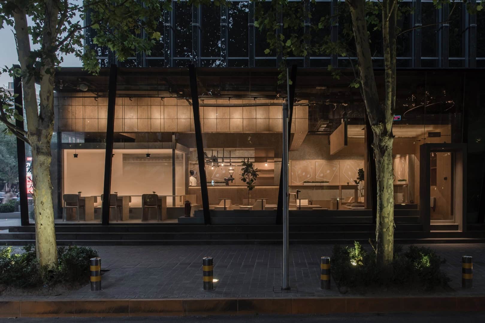 Modern glass commercial building with illuminated interior, sleek architecture, and surrounding trees at dusk.
