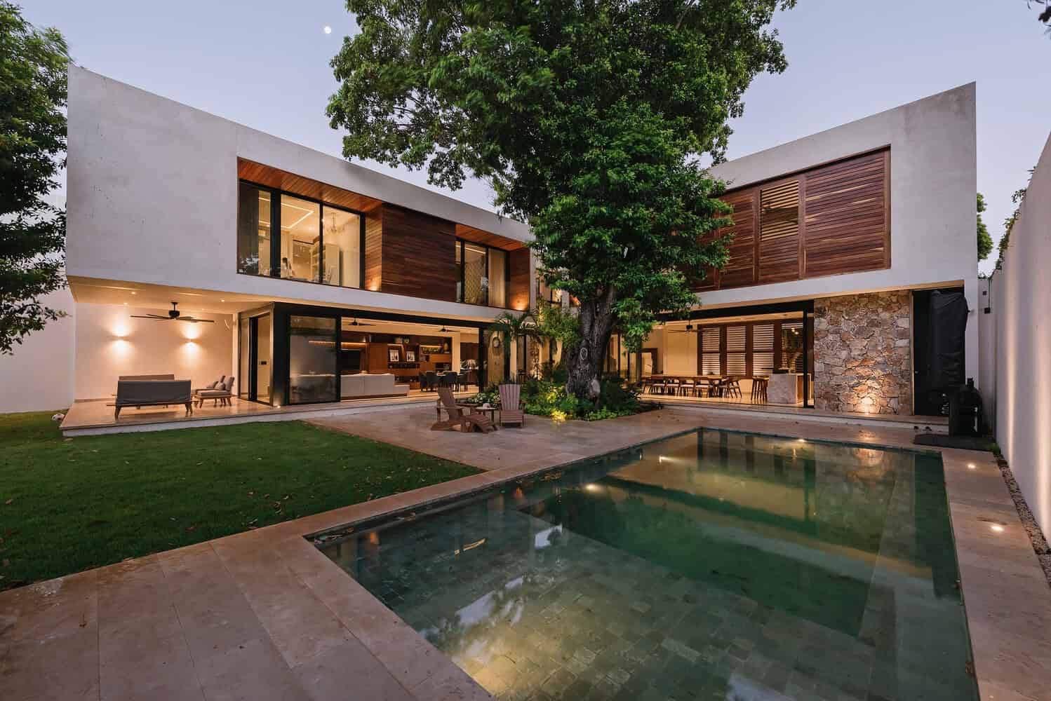 Contemporary modern house with sleek architecture, wooden accents, large glass windows, and a luxurious pool in a landscaped garden at dusk.