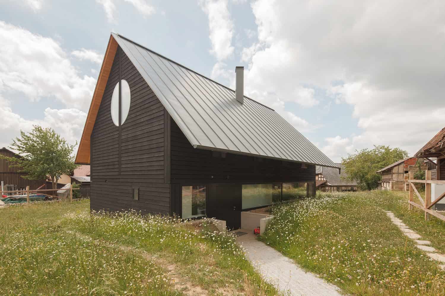 Contemporary black wooden house with a steep metal roof and circular window, set in a lush grassy landscape with wildflowers, under a partly cloudy sky, showcasing modern architecture and innovative home design.