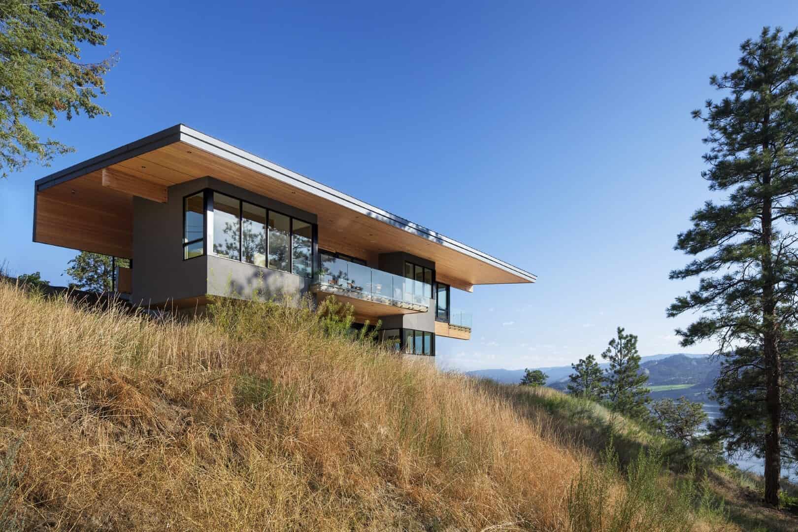 Modern hillside house with extensive glass windows, wooden accents, and a flat roof, set against a scenic mountain landscape for contemporary architecture and luxury home design.