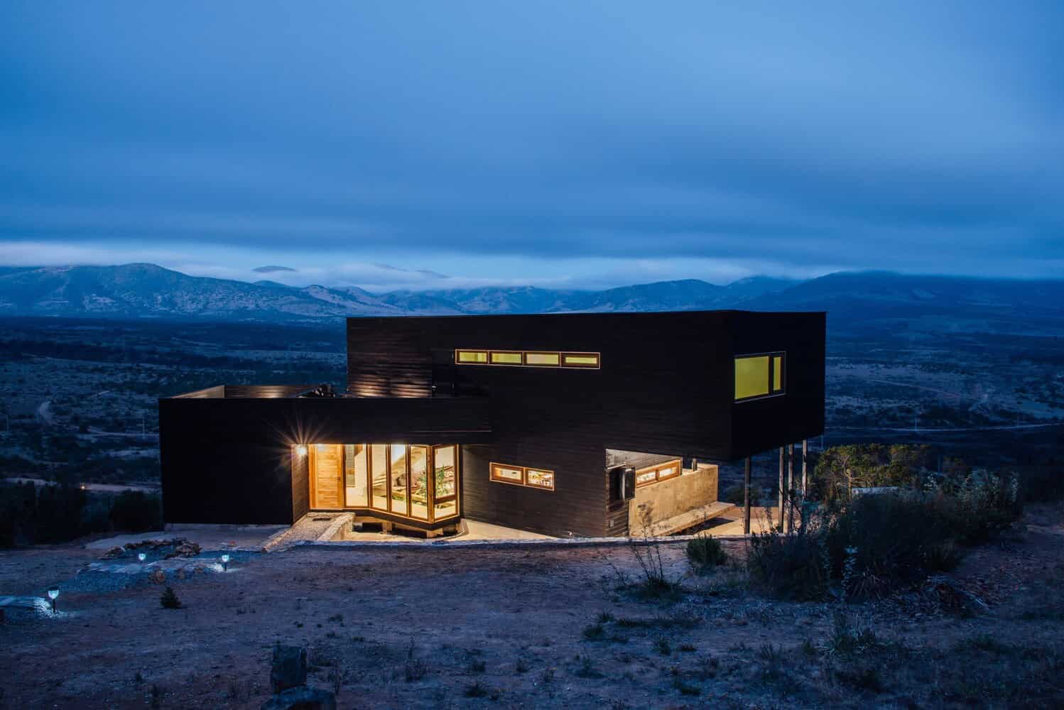 Modern black minimalist house with large windows and contemporary architecture set against mountainous landscape at dusk.