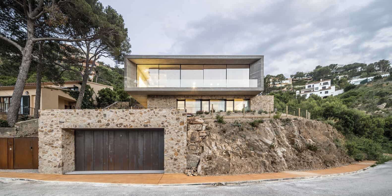 Luxury modern hillside house with large glass windows and stone facade, blending contemporary architecture with natural landscape, in a scenic mountain setting.