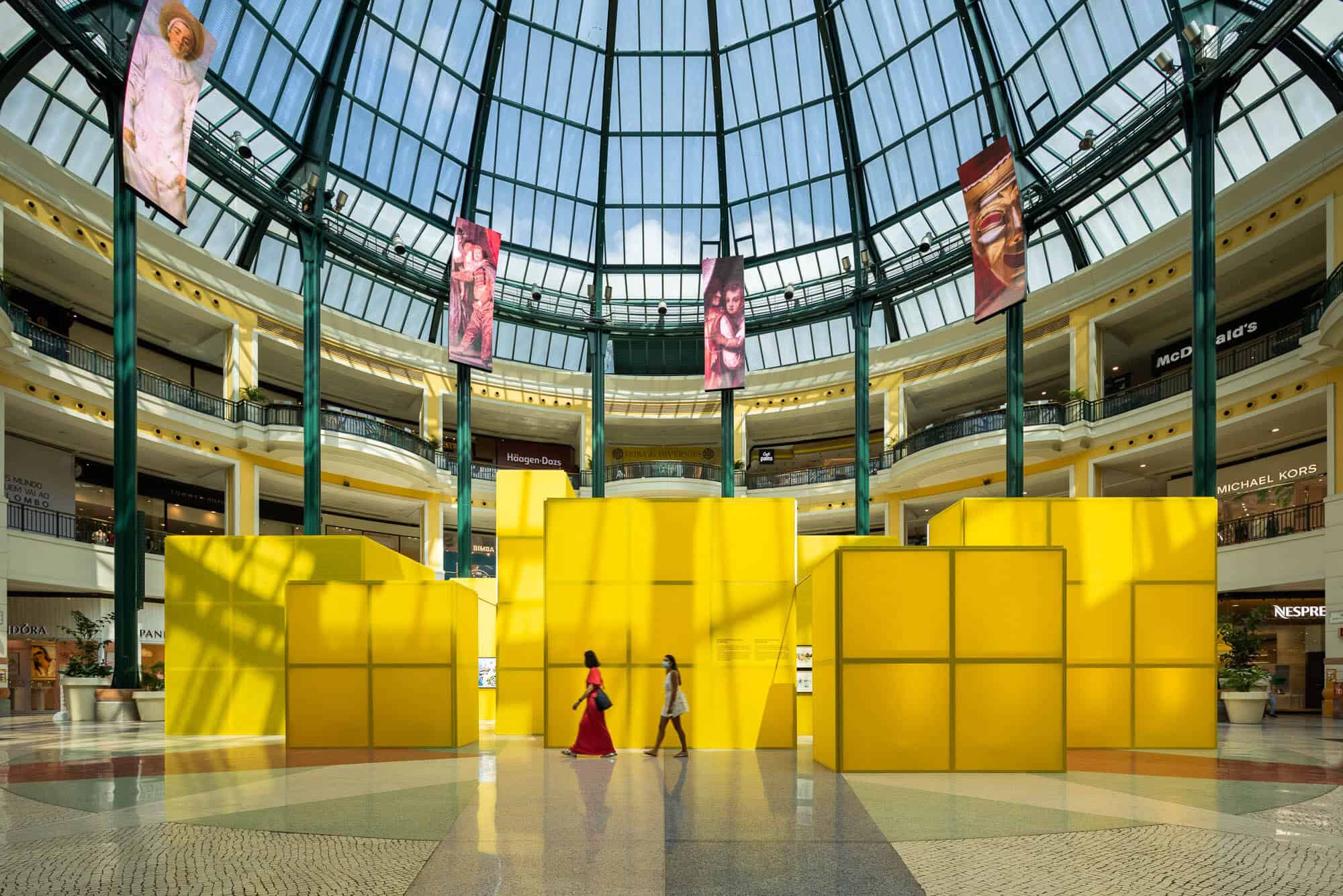 Vibrant yellow art installation inside a modern shopping mall with glass domed roof, showcasing contemporary architecture and innovative interior design elements.